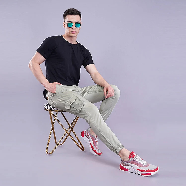 Meesho seller photoshoot example of an Indian male model in a black crew-neck T-shirt, beige cargo joggers and white-red sneakers, seated on a folding stool wearing tinted sunglasses against a soft lavender studio backdrop