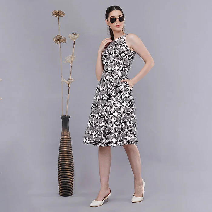 Indian female model in a black and white geometric print sleeveless knee-length dress with sunglasses and white heels, standing full length against a soft grey studio backdrop beside a tall vase, a clean on-model image ready for amazon a+ content for fashion brands