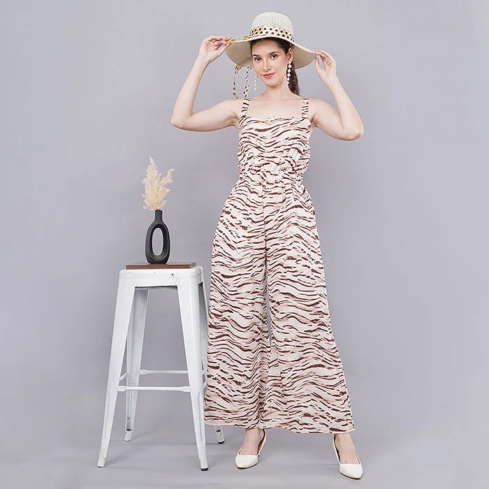 Full-length studio fashion photo of an Indian female model in a sleeveless square-neck jumpsuit with a beige and brown zebra print, wide-leg trousers, a straw hat and nude heels beside a white stool and vase on a light grey backdrop, showing neckline, sleeve and length attributes clearly