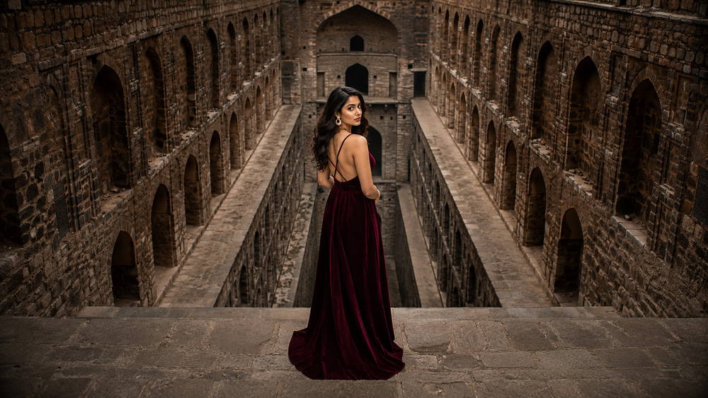 Dramatic cinematic editorial photograph of a female Indian model in deep burgundy velvet dress standing at the entrance of Agrasen Ki Baoli stepwell Delhi with symmetrical descending steps forming striking geometric backdrop and moody shadows