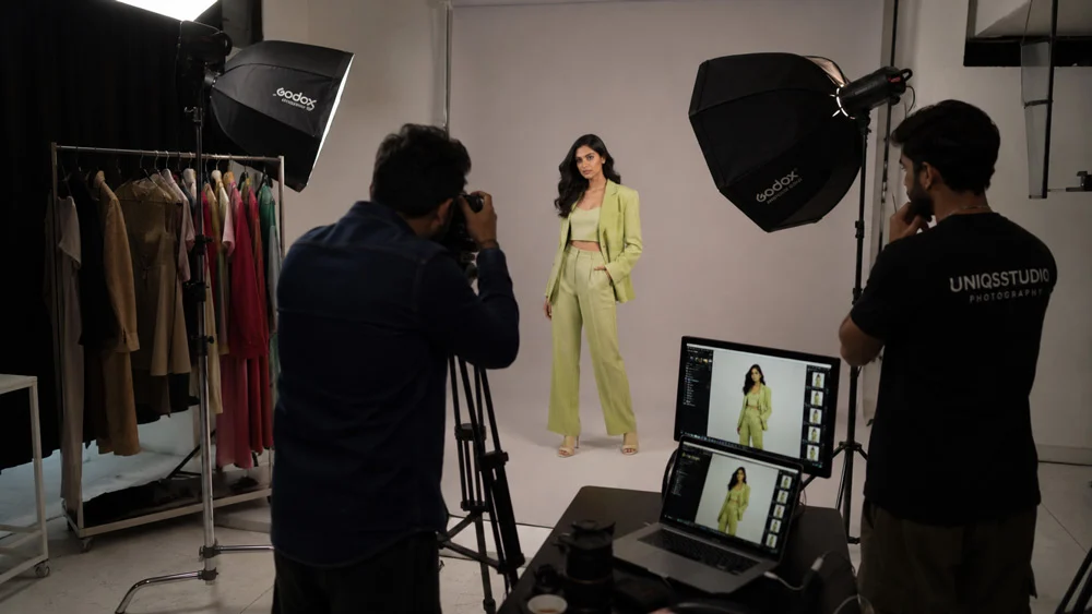 Behind the scenes of a fashion photography shoot in an Indian studio: a photographer and assistant shoot a model in a lime green pantsuit under a large octabox, with a clothing rack and a tethered monitor showing the frames