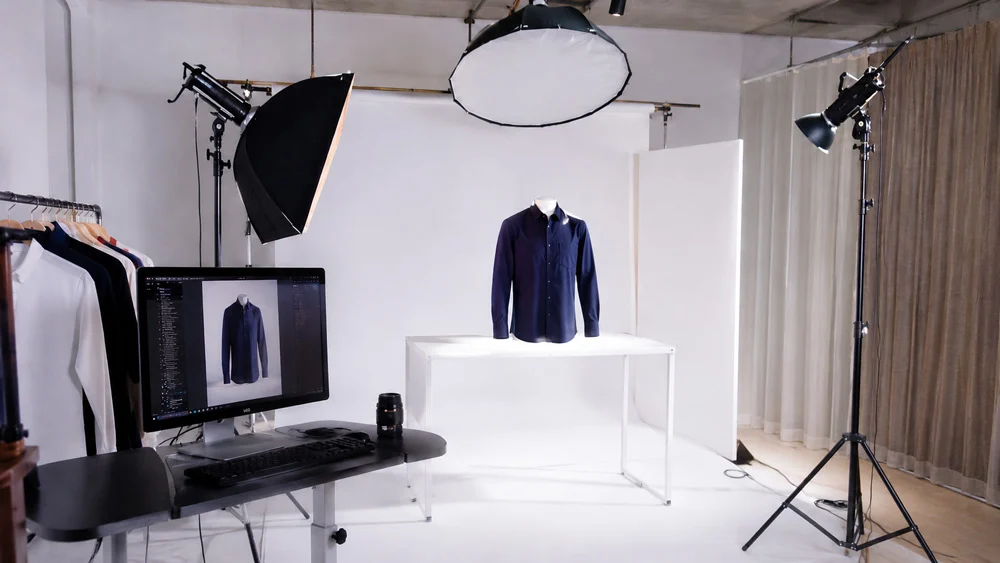 Ghost mannequin photography studio setup at Uniqs Studio Delhi with a navy shirt on an invisible mannequin on a white table, octabox and strip softboxes, a tethered monitor showing the garment and a clothing rack, the kind of consistent catalogue setup that helps Meesho sellers cut returns