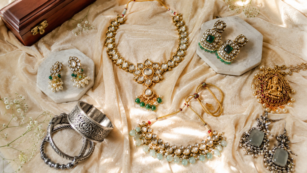 Overhead flat lay of assorted Indian fashion jewellery including Kundan necklace Polki earrings temple gold pendant and oxidized silver bangles arranged on cream velvet cloth with soft window light