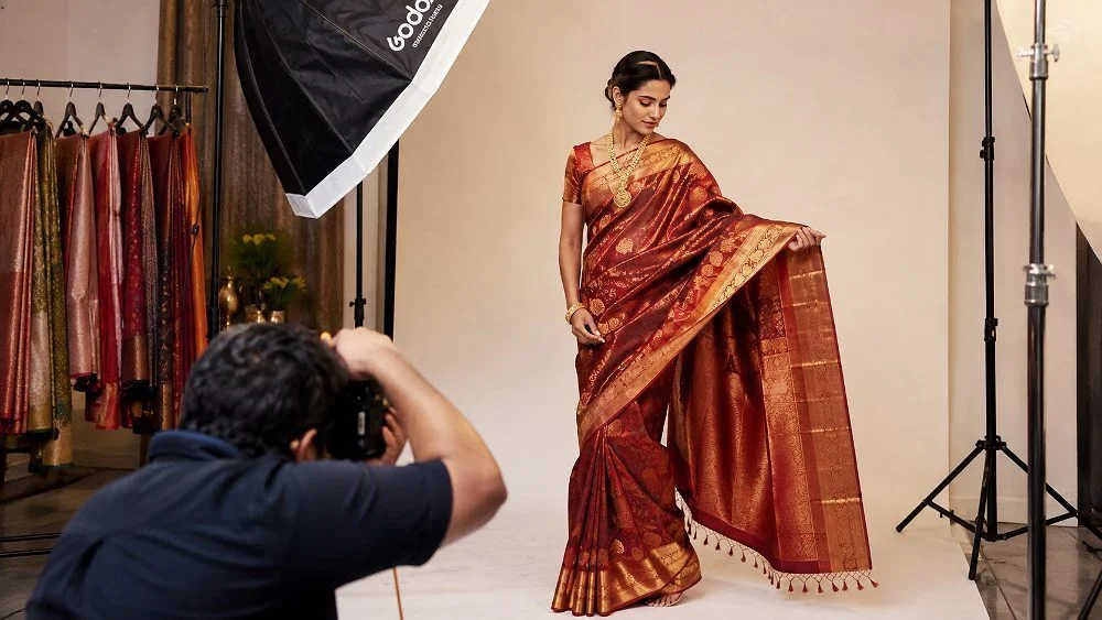 Behind-the-scenes meesho product photography of an Indian model draped in a maroon and gold silk saree, photographer crouching with a mirrorless camera under a large softbox, rack of colourful sarees in a warm-toned ecommerce studio