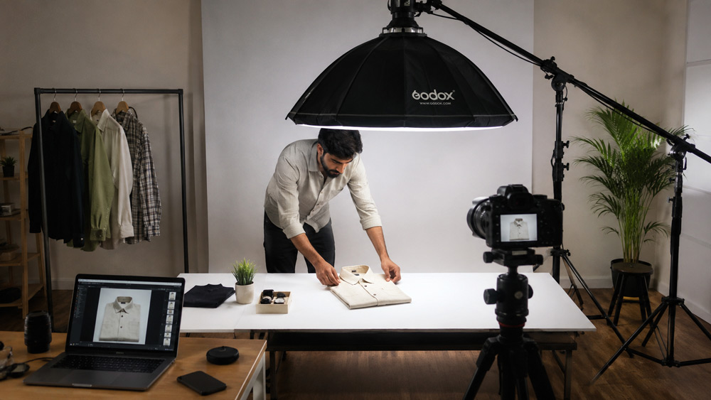 Behind the scenes of professional garment flat lay photography shoot at Uniqs Studio Delhi NCR with overhead tripod softbox lighting stylist arranging folded shirt on white background modern minimal studio interior editorial documentary style