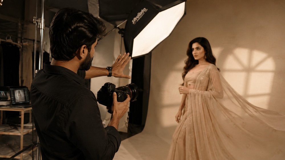 Editorial magazine-style photograph of a professional Indian model photographer holding a DSLR camera, directing a female fashion model in a soft-flowing beige outfit inside a premium modern studio in Delhi NCR with cinematic softbox lighting and champagne-gold color grade