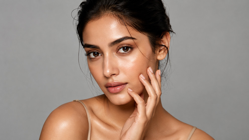 Clean beauty headshot for Model Photography Portfolio — Indian female model with flawless dewy skin, natural nude makeup, direct camera gaze, shot on 85mm lens with soft continuous lighting on clean grey seamless backdrop, editorial magazine-cover quality