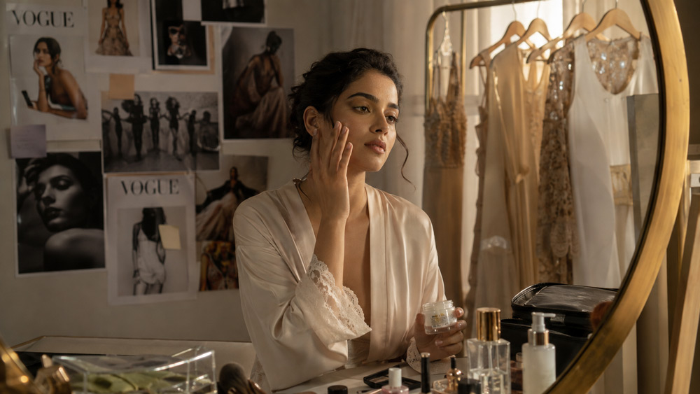 Editorial fashion photograph of a young Indian female model in her mid-20s sitting at a vanity in a luxury photography studio in Delhi, softly lit by warm natural window light, applying subtle skincare, wardrobe rack and mood board behind her, ivory and champagne gold palette, Vogue magazine aesthetic